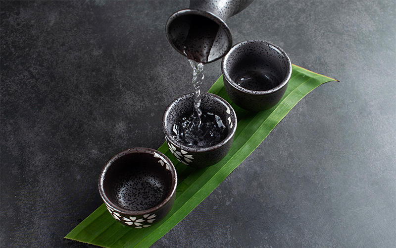 clear-chinese-liquor-being-poured-from-ceramic-carafe-into-traditional-tasting-cups-over-green-leaf